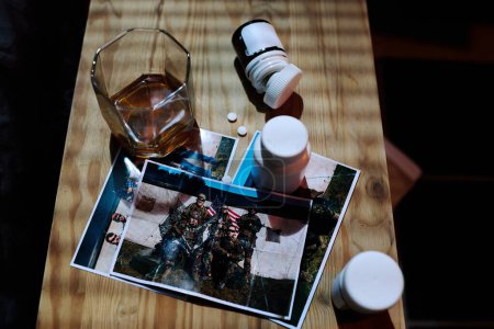 Photo for Above angle of wooden night table with bottles with various pills, photos of army friends and glass of whisky standing by bed - Royalty Free Image
