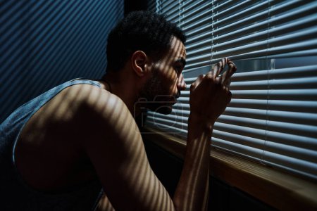 Photo for Side view view of young sleepless man looking through venetian blinds on window during sneak peek after someone outdoors - Royalty Free Image