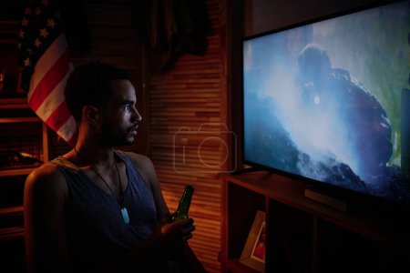 Photo for Young sleepless man with bottle of beer or other alcohol watching video on screen of tv set about his taking part in military operation - Royalty Free Image