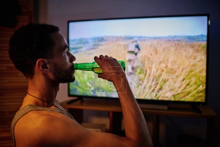 Photo for Young man with post traumatic disorder having beer while watching video about military operation where he took part - Royalty Free Image