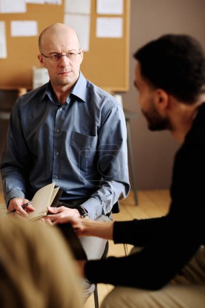 Confident mature counselor looking at one of patients with post traumatic disorder while listening to him during session