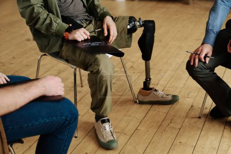 Foto de Primer plano de las piernas del joven con discapacidad física sentado en la silla entre otros hombres durante la discusión de sus problemas psicológicos - Imagen libre de derechos