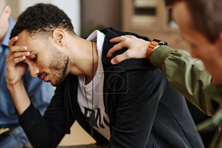 Young stressed man touching head while one of attendants keeping hand on his shoulder and supporting him during psychological session