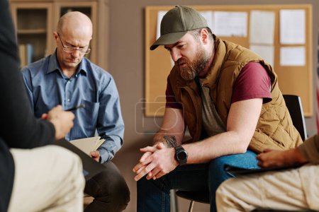 Photo for Young upset man describing traumatic event or experience from his life while sitting among counselor and other attendants of session - Royalty Free Image