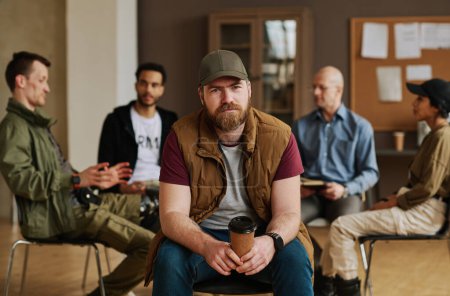 Téléchargez les photos : Homme barbu avec une tasse de café regardant la caméra tout en étant assis contre un groupe de personnes décrivant leurs problèmes psychologiques - en image libre de droit