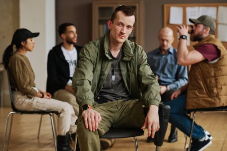 Hombre joven caucásico con síndrome postraumático sentado frente a la cámara en el fondo de personas interculturales del grupo de apoyo