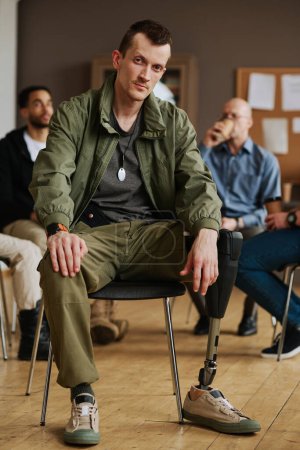 Photo for Young man with prosthetic leg sitting in front of camera against counselor and other attendants of PTSD support group having break - Royalty Free Image