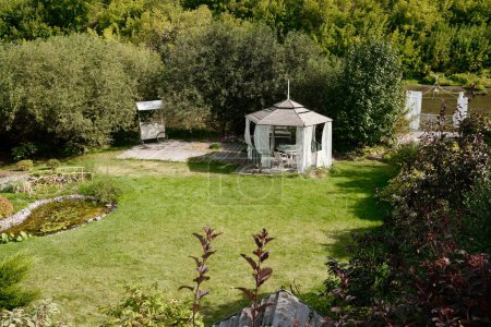 Téléchargez les photos : Grande pelouse verte ensoleillée sur la cour de la maison d'été avec alcôve entourée d'un petit étang, de buissons et d'arbres - en image libre de droit
