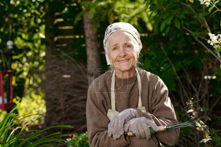 Photo for Happy senior woman with small rake for loosening soil looking at camera while standing in the garden with green plants on sunny day - Royalty Free Image