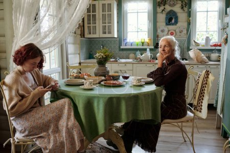 Téléchargez les photos : Grand-mère reposante en tenue décontractée intelligente assise à côté de la table devant sa petite-fille défilant dans le smartphone dans la cuisine - en image libre de droit