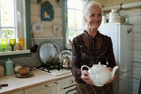 Téléchargez les photos : Grand-mère souriante en tenue décontractée intelligente portant une théière en porcelaine blanche avec du thé dans le salon où ses invités attendent le petit déjeuner - en image libre de droit