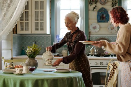 Téléchargez les photos : Grand-mère verser une tisane fraîche dans des tasses tandis que la jeune fille portant tarte maison pour le mettre sur la table servi pour le petit déjeuner - en image libre de droit