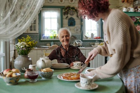 Téléchargez les photos : Heureuse femme âgée assise près de la table servie et regardant sa petite-fille couper une tarte sucrée maison fraîche - en image libre de droit