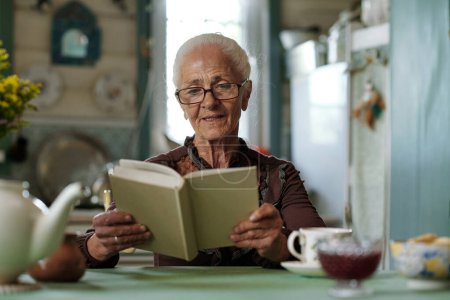 Photo for Senior woman in eyeglasses reading captivating story or novel after breakfast while sitting by kitchen table in summer house - Royalty Free Image