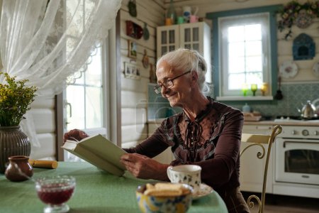 Photo for Happy aged woman in smart casualwear and eyeglasses reading novel or book of stories while sitting by table in the kitchen of summer house - Royalty Free Image