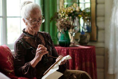 Téléchargez les photos : Jolie femme âgée avec toile en cerceau coutures de croix à loisir tout en étant assis sur le canapé dans le salon de la maison de campagne - en image libre de droit