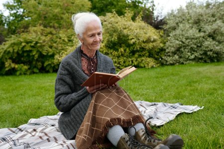 Photo for Senior woman in casualwear sitting on checkered woolen plaid on green lawn and reading book while having rest by summer house - Royalty Free Image