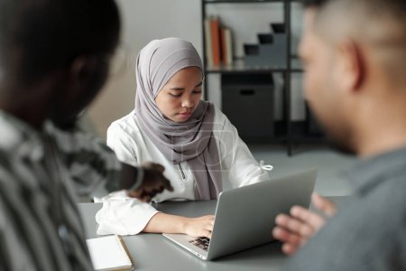 Foto de Joven estudiante musulmana trabajando en red por escritorio mientras dos crueles hombres interculturales la señalan y se burlan de ella - Imagen libre de derechos