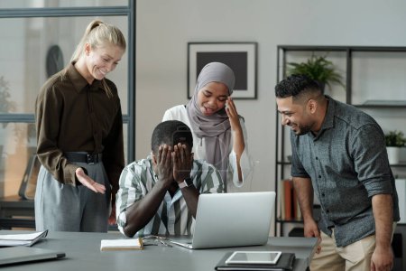 Téléchargez les photos : Groupe de jeunes étudiants multiculturels riant de l'homme noir cachant son visage tout en pleurant par le lieu de travail entouré de camarades de classe cruels - en image libre de droit