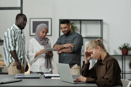Foto de Joven mujer de negocios rubia tocando la cabeza mientras está sentado en el lugar de trabajo frente a la computadora portátil contra el grupo de colegas interculturales - Imagen libre de derechos