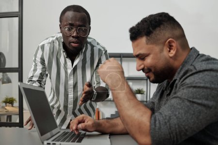 Foto de Joven jefe afroamericano enojado o gerente regañando a colega masculino hispano sentado frente a la computadora portátil y tratando de terminar el trabajo - Imagen libre de derechos