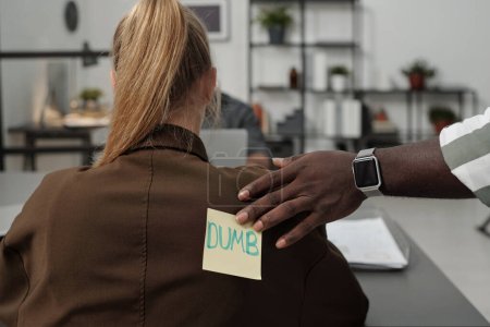 Foto de African American businessman putting sticker with word dumb on back of blond female colleague of another ethnicity while making fun of her - Imagen libre de derechos