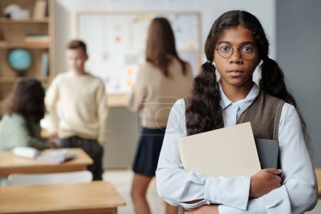 Photo for Serious secondary school learner of African ethnicity looking at camera while standing against group of classmates chatting at break - Royalty Free Image