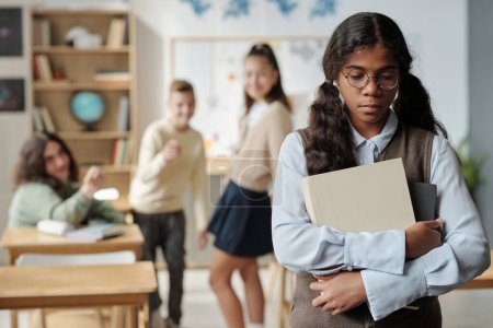 Photo for Pre-teen upset schoolgirl with books standing in front of camera with offended expression against three classmates bullying her - Royalty Free Image