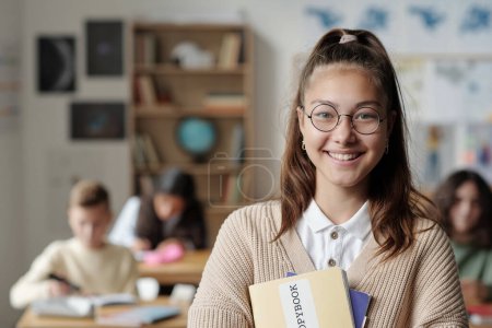 Foto de Sonriente colegiala joven en gafas y ropa casual mirando a la cámara mientras está de pie delante de sus compañeros de clase en la lección - Imagen libre de derechos