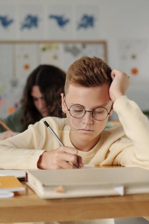 Photo for Serious pre-teen schoolboy in eyeglasses making notes in copybook during individual assignment at lesson against his classmate - Royalty Free Image