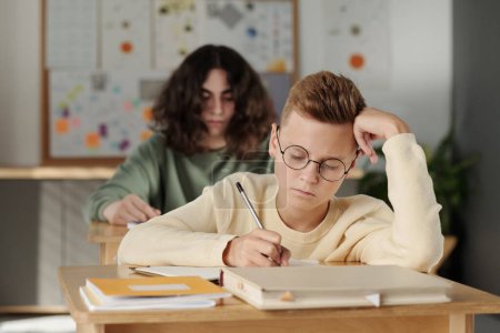 Téléchargez les photos : Écolier intelligent jeune dans les lunettes de prendre des notes dans le copybook lors d'une affectation individuelle à la leçon contre son camarade de classe - en image libre de droit