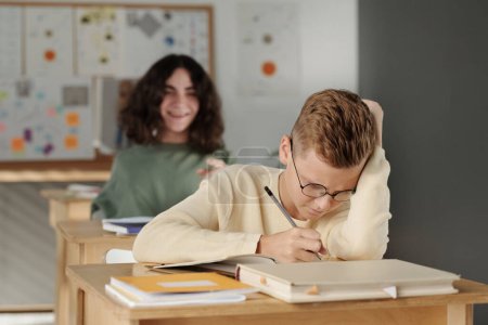 Photo for Selective focus on clever and diligent schoolboy making notes in copybook at lesson against cruel classmate laughing at him - Royalty Free Image