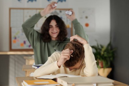 Téléchargez les photos : Jeune écolier se penchant sur le bureau et se couvrant la tête par les mains tandis que son camarade de classe lui jetant du papier froissé à la leçon - en image libre de droit