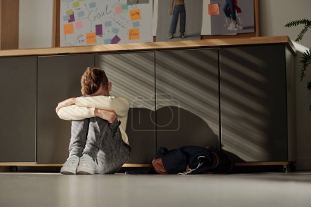 Photo for Helpless secondary school pupil sitting on the floor of classroom and hiding his head in arms while suffering from bullying - Royalty Free Image