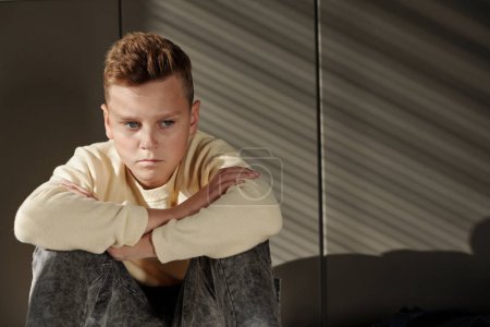 Photo for Gloomy schoolboy in casualwear looking aside while sitting on the floor after lessons and suffering from bullying of his classmates - Royalty Free Image