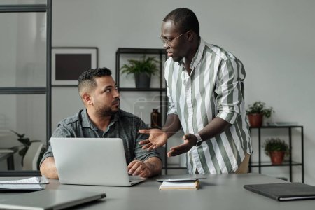 Young irritated African American employee looking at multi-ethnic or Hispanic man sitting by desk in front of laptop during conversation
