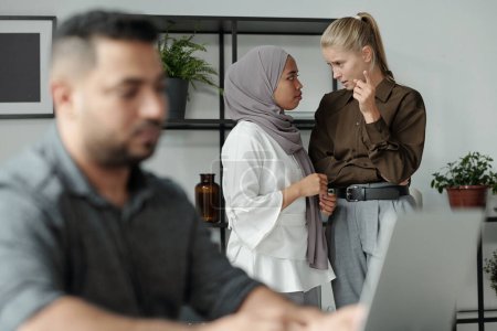 Photo for Young blond businesswoman discussing male colleague of another ethnicity with black woman in hijab while standing behind him - Royalty Free Image
