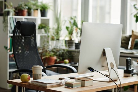 Foto de Lugar de trabajo del trabajador moderno de cuello blanco con monitor de computadora, manzana fresca y taza de café y sillón negro en el espacio de coworking - Imagen libre de derechos
