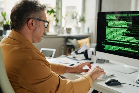 Téléchargez les photos : Vue latérale d'un ingénieur informatique masculin sérieux mature tapant sur un clavier d'ordinateur assis devant un écran avec des données codées - en image libre de droit