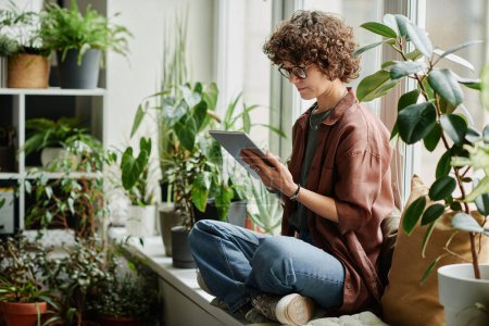 Téléchargez les photos : Jeune jolie femme brune avec tablette numérique assise sur le rebord de la fenêtre parmi une variété de plantes de serre et de réseautage - en image libre de droit