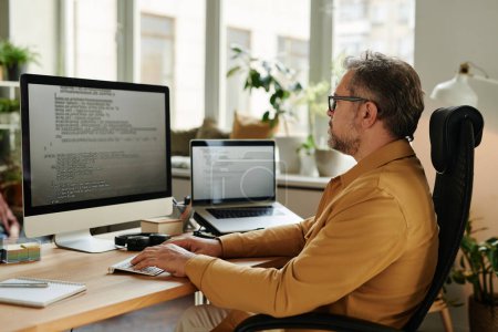 Foto de Desarrollador de software maduro serio que mira el código en la pantalla del ordenador mientras decodifica datos y realiza tareas de trabajo en la oficina - Imagen libre de derechos