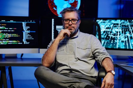 Téléchargez les photos : Développeur de logiciels masculins mûrs sérieux en vêtements décontractés intelligents et lunettes assis contre des écrans d'ordinateur dans la salle de serveur - en image libre de droit