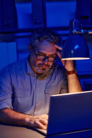 Foto de Programador masculino maduro cansado sentado por el lugar de trabajo delante de la computadora portátil en la oficina y encontrar maneras de decodificar datos mientras trabaja horas extras - Imagen libre de derechos