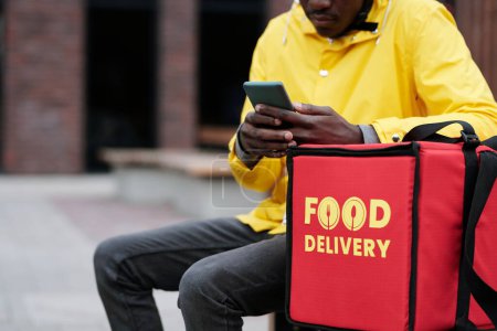 Téléchargez les photos : Plan recadré d'un jeune livreur assis sur un banc par un gros sac rouge et vérifiant de nouvelles commandes en ligne de nourriture sur un téléphone portable à l'extérieur - en image libre de droit