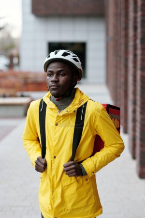 Téléchargez les photos : Jeune homme noir sérieux avec casque de sécurité et veste jaune avec sac à dos sur le dos debout devant la caméra en milieu urbain - en image libre de droit