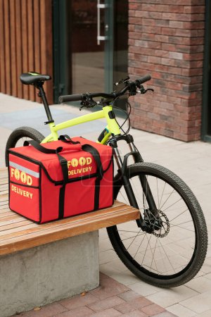 Téléchargez les photos : Bicyclette de messagerie garée par banc en bois avec grand sac à dos carré rouge avec nourriture commandée pour les clients en ligne du service de livraison - en image libre de droit
