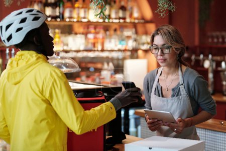 Téléchargez les photos : Jeune serveuse en tablier regardant la liste des commandes à l'écran de smartphone tenue par un courrier afro-américain en casque et veste jaune - en image libre de droit