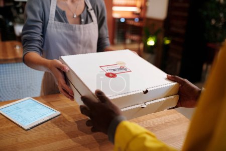 Photo for Hands of young female worker of cafe passing two packed square boxes of pizza to courier over wooden counter while preparing orders - Royalty Free Image