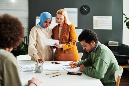 Téléchargez les photos : Mature professeur blond montrant le document avec un test de grammaire anglaise à une étudiante musulmane dans le hijab tout en étant debout par le lieu de travail - en image libre de droit