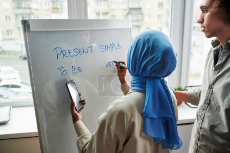 Foto de Vista posterior de una joven profesora musulmana explicando a un chico multiétnico Presente Simple mientras escribe ejemplos en pizarra - Imagen libre de derechos